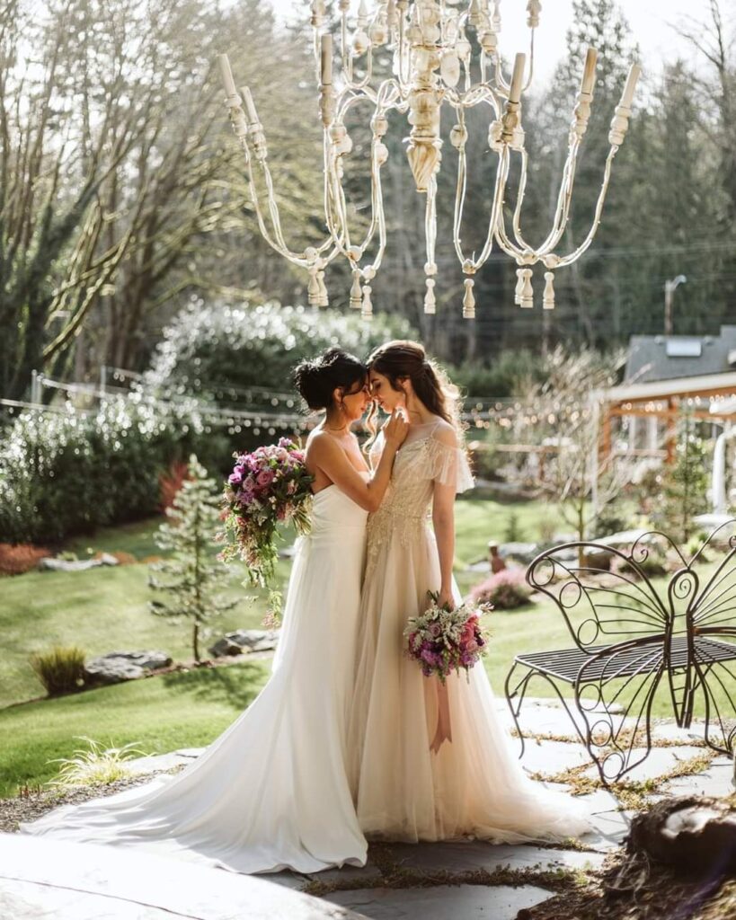 Two brides in white gowns holding floral bouquets foreheads touching under an outdoor chandelier in a garden setting