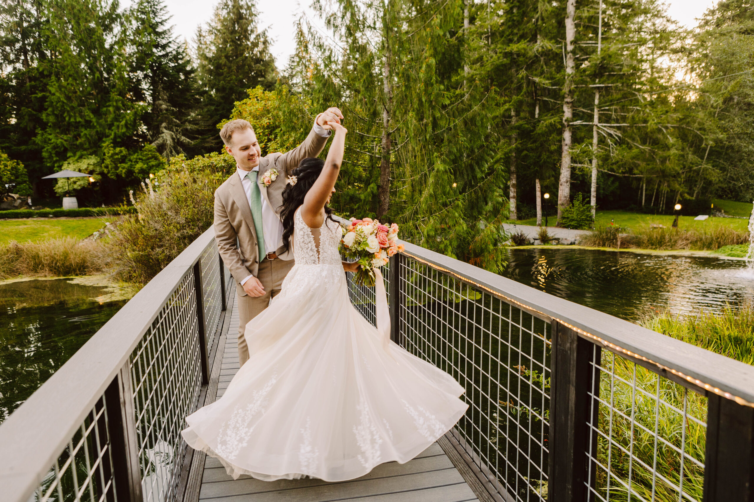 Bride in a white wedding gown and a tan suit groom dancing on a wooden bridge over a pond surrounded by trees