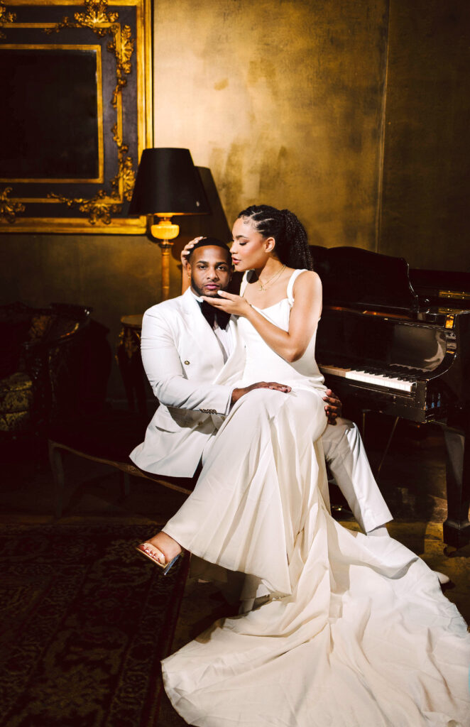 Bride in white gown sitting on grooms lap in white tuxedo beside a grand piano in a gold toned elegant room