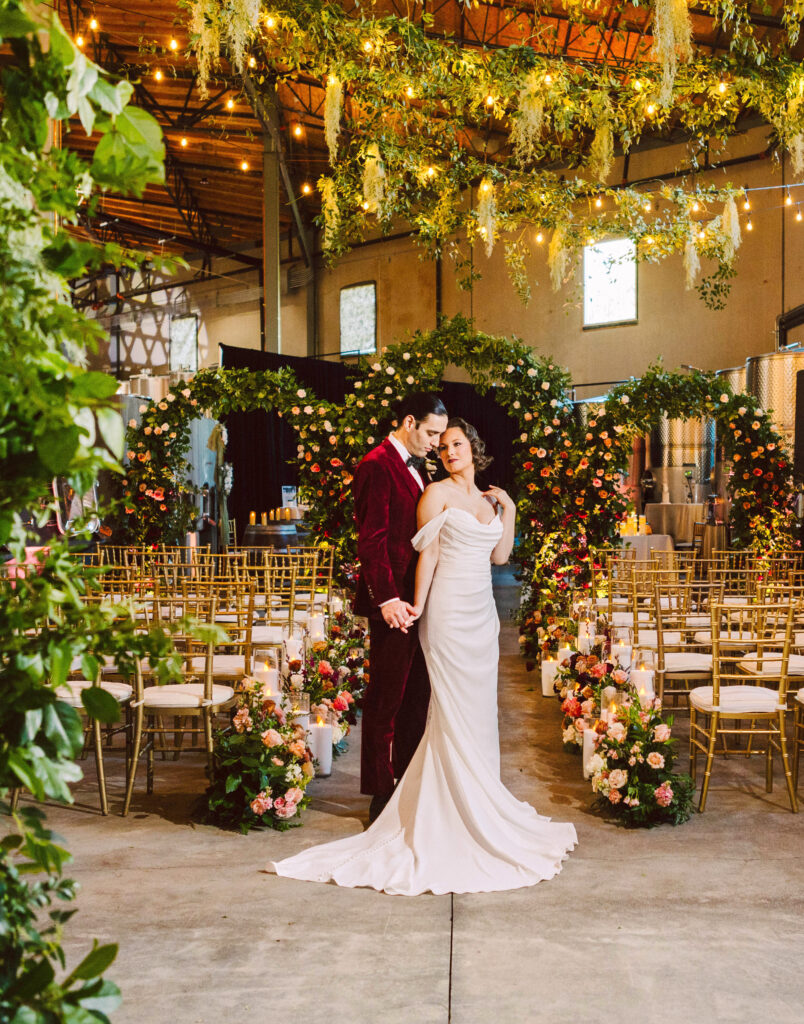 Couple in wedding attire standing together in a flower lined aisle inside a rustic venue with hanging greenery and string lights