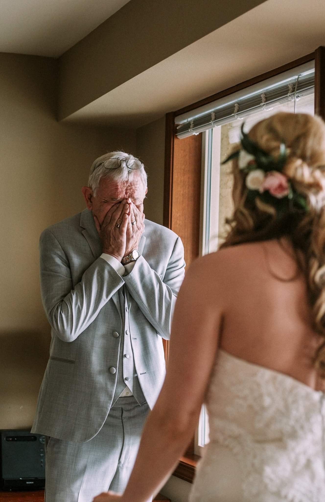 Older man in a light gray suit covers his face with his hands standing by a window at a wedding as the bride in a lace dress is seen from behind in the foreground