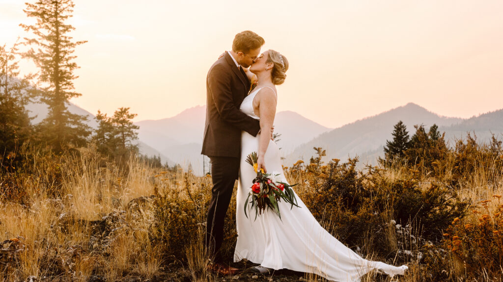 A bride and groom share a kiss in a sunlit grassy field with mountains in the distance and warm sunset tones the bride holds a bouquet