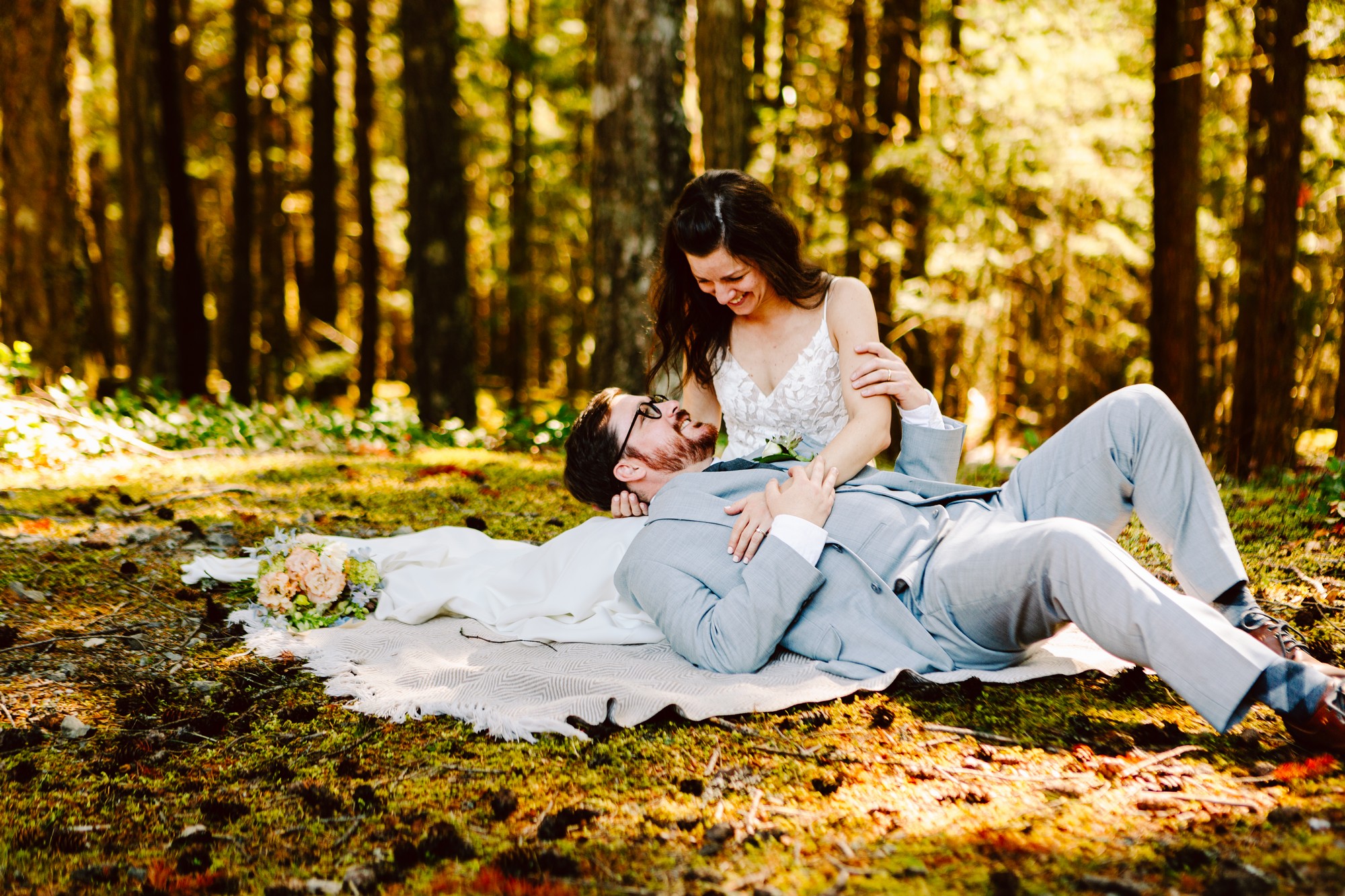 Bride in white dress and groom in light gray suit laughing together on a blanket in a sunlit forest.