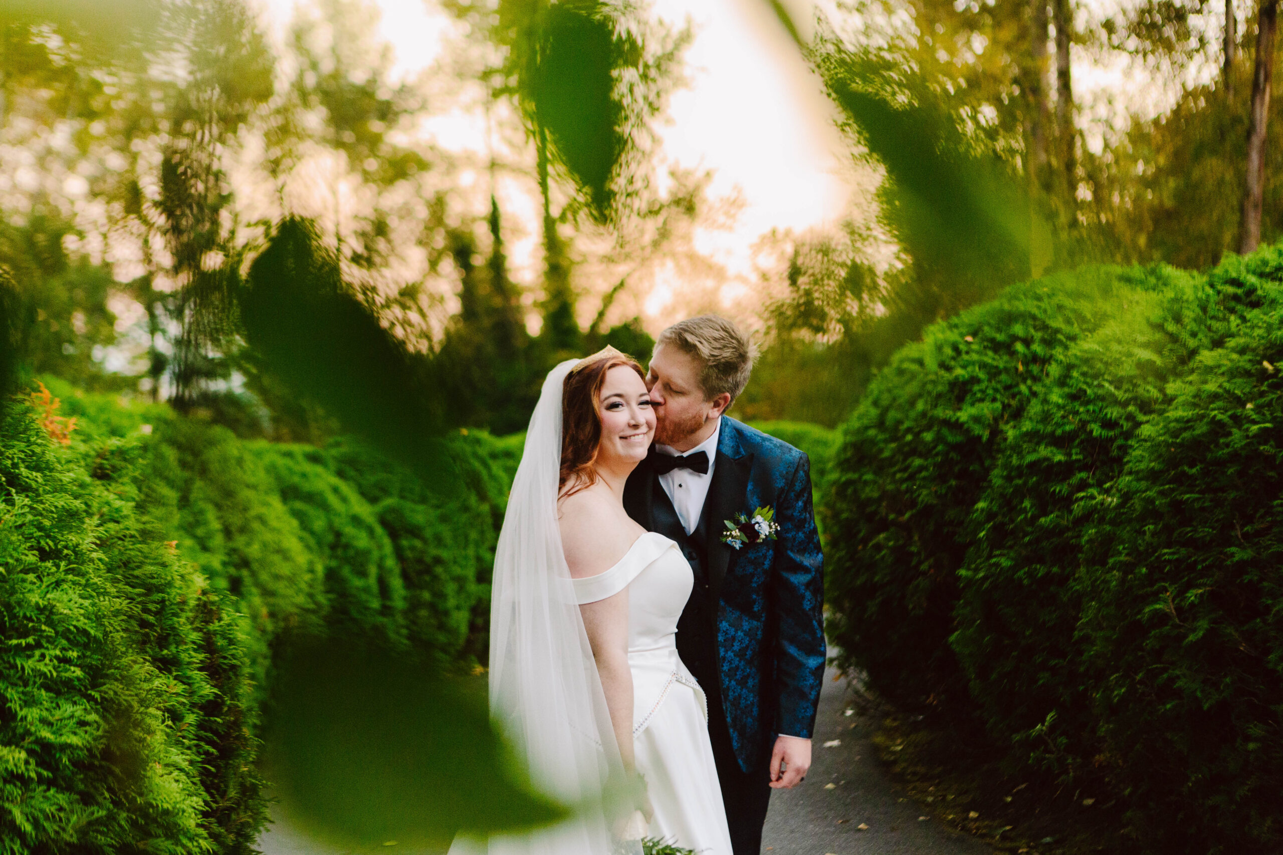 Bride in a white wedding gown with veil stands beside a groom in a blue suit, along a lush green garden path, smiling at the camera.