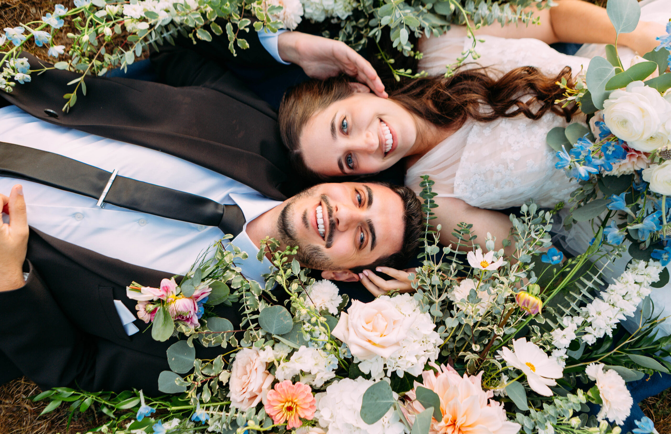 Bride and groom lying on the ground surrounded by lush floral arrangements of pink, white, and blue flowers with eucalyptus.