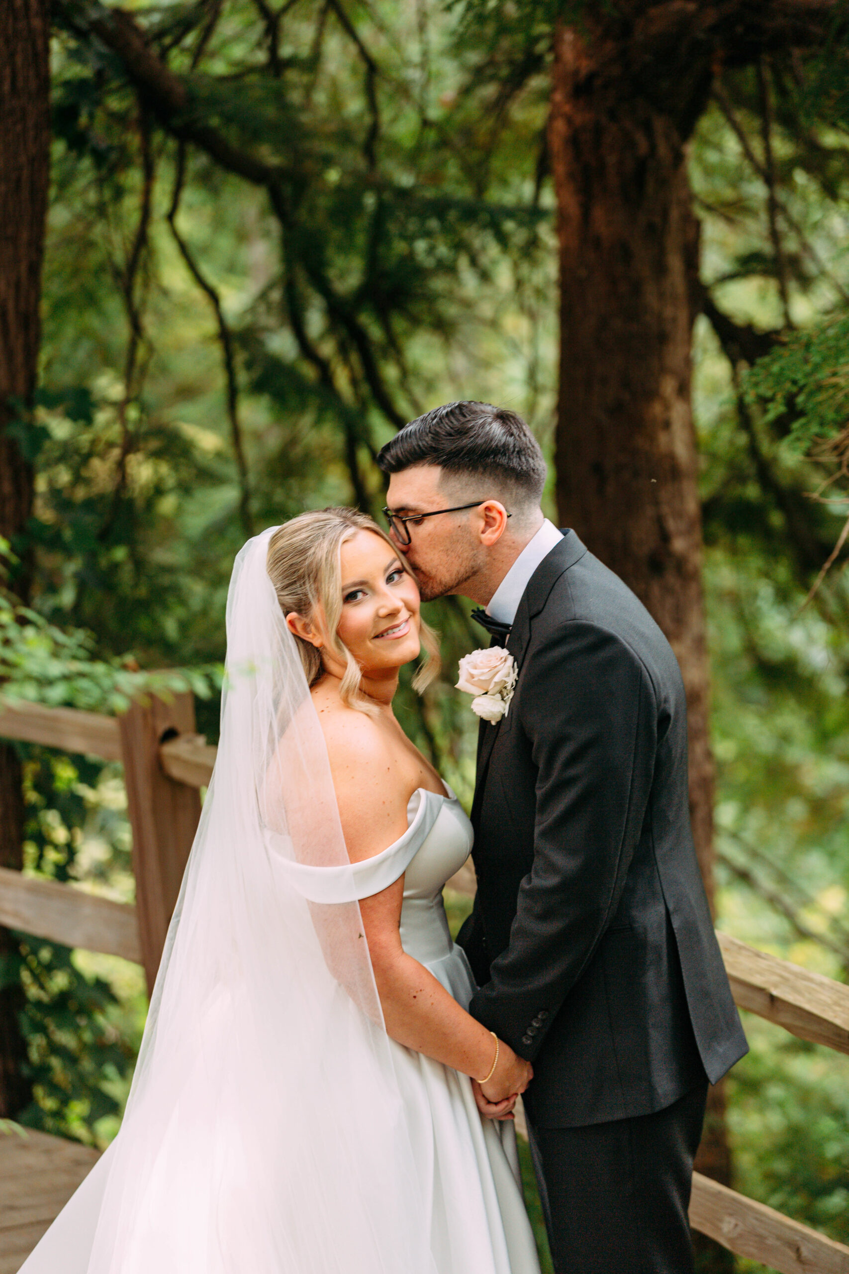 Groom in dark suit kissing bride's cheek as she smiles, standing on a wooden bridge surrounded by lush green forest.