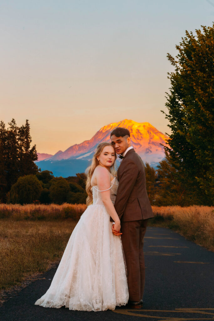 Bride and groom embrace on a road at sunset with a glowing mountain in the background