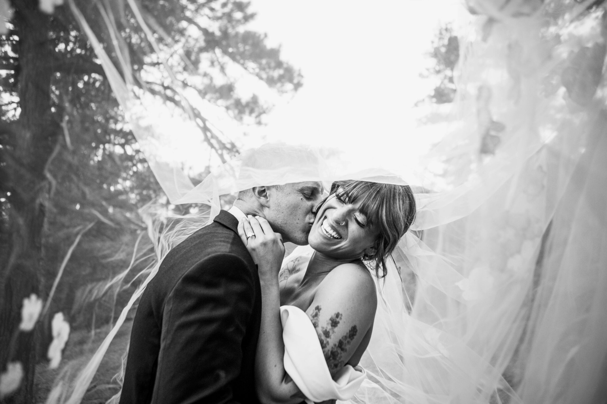 Groom kissing smiling bride on the cheek beneath a flowing veil, black and white outdoor wedding portrait among trees.