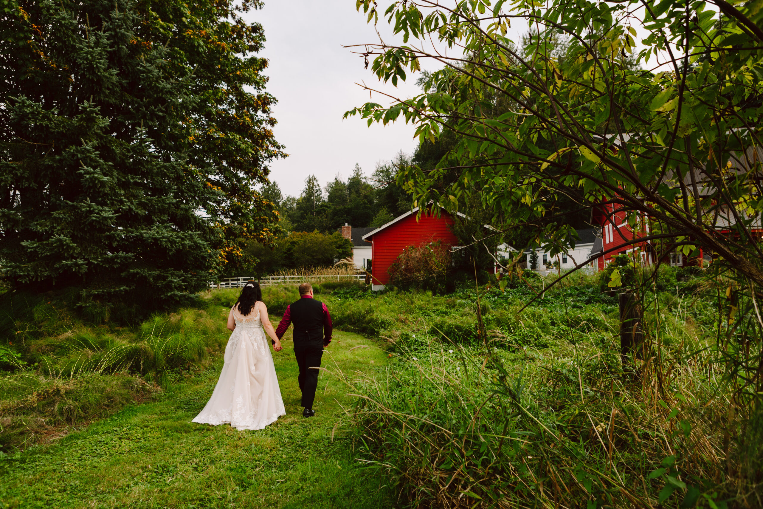 Bride and groom walking hand-in-hand through a lush green path toward a red barn surrounded by trees.