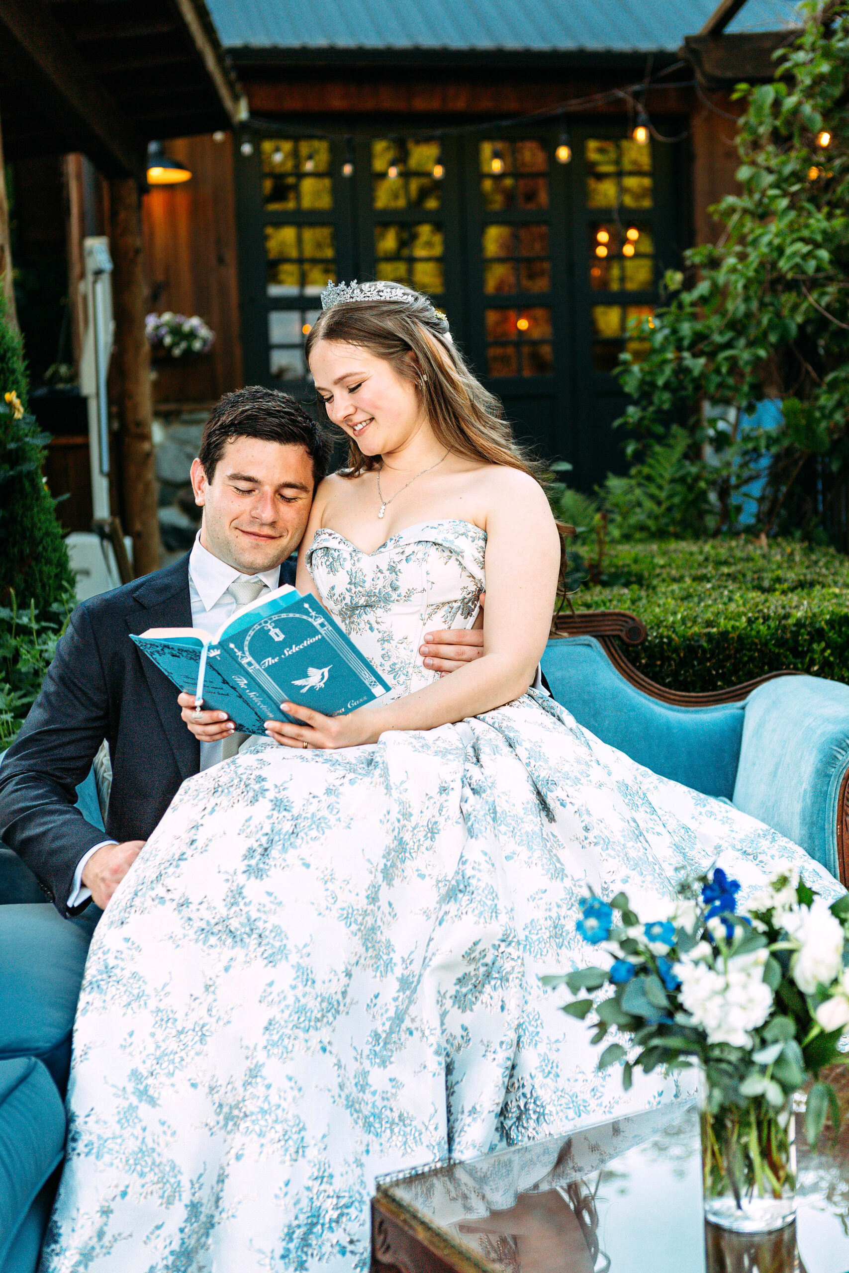 Bride in floral gown and tiara reading "The Selection" with groom on a blue sofa outside a rustic venue with string lights.