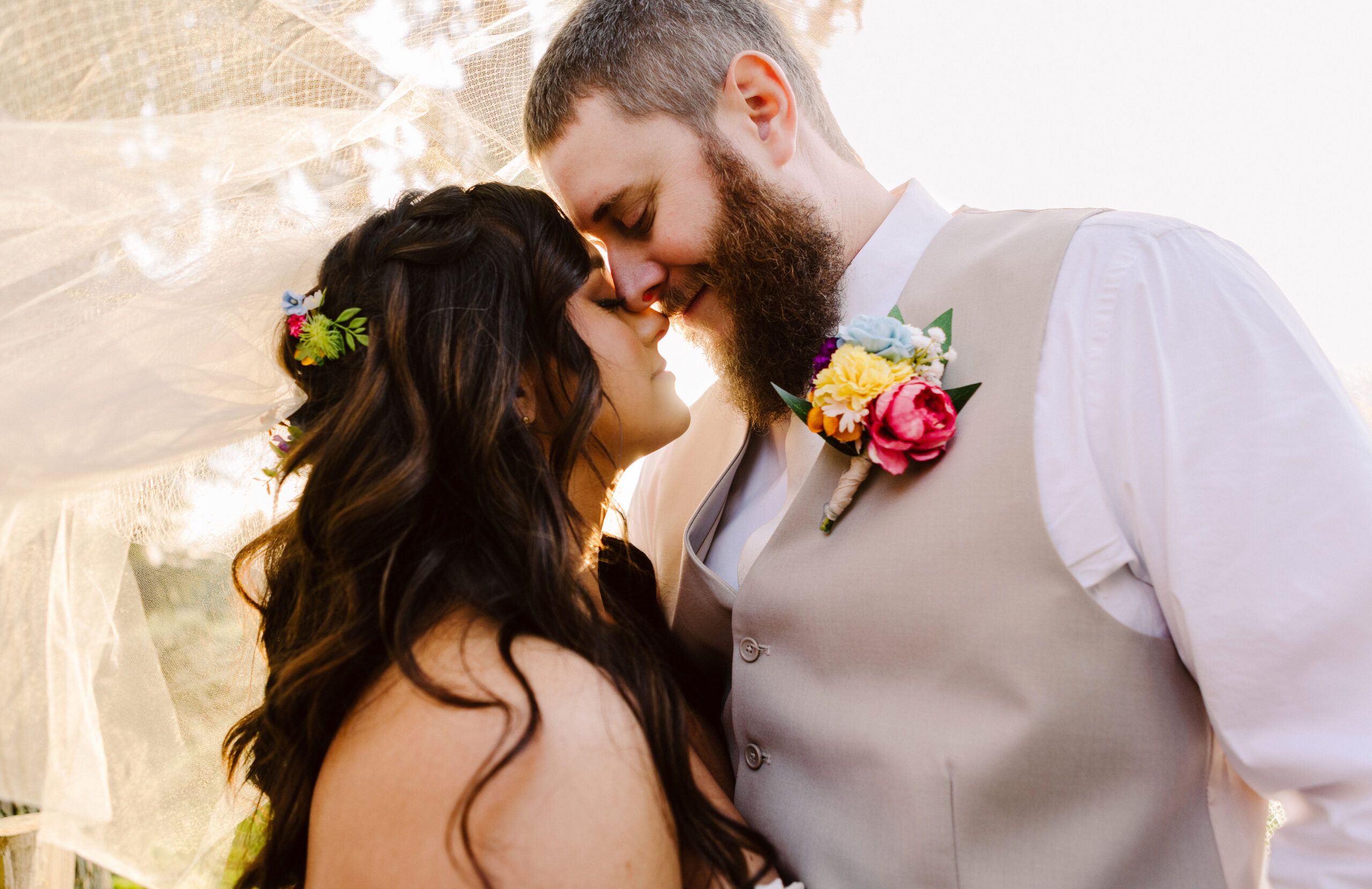 Bride and groom touching foreheads under a veil, she wears a floral hair piece, he wears a colorful boutonnière on a tan vest.