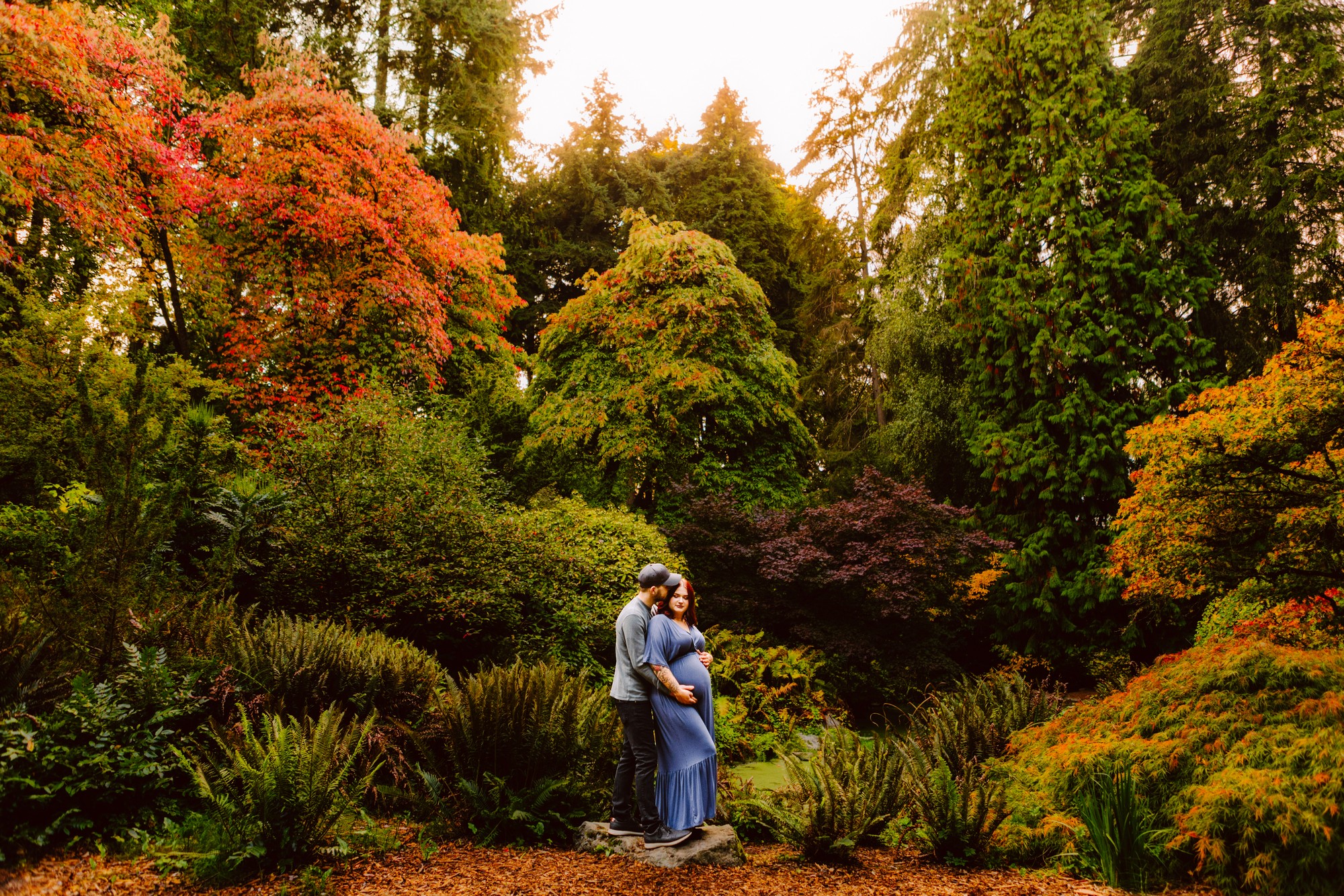Pregnant woman and partner standing together in a lush autumn garden with colorful trees and leaves on the ground in fall