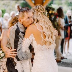 Bride in lace wedding dress with floral hair piece embracing an elderly man at an outdoor wedding venue
