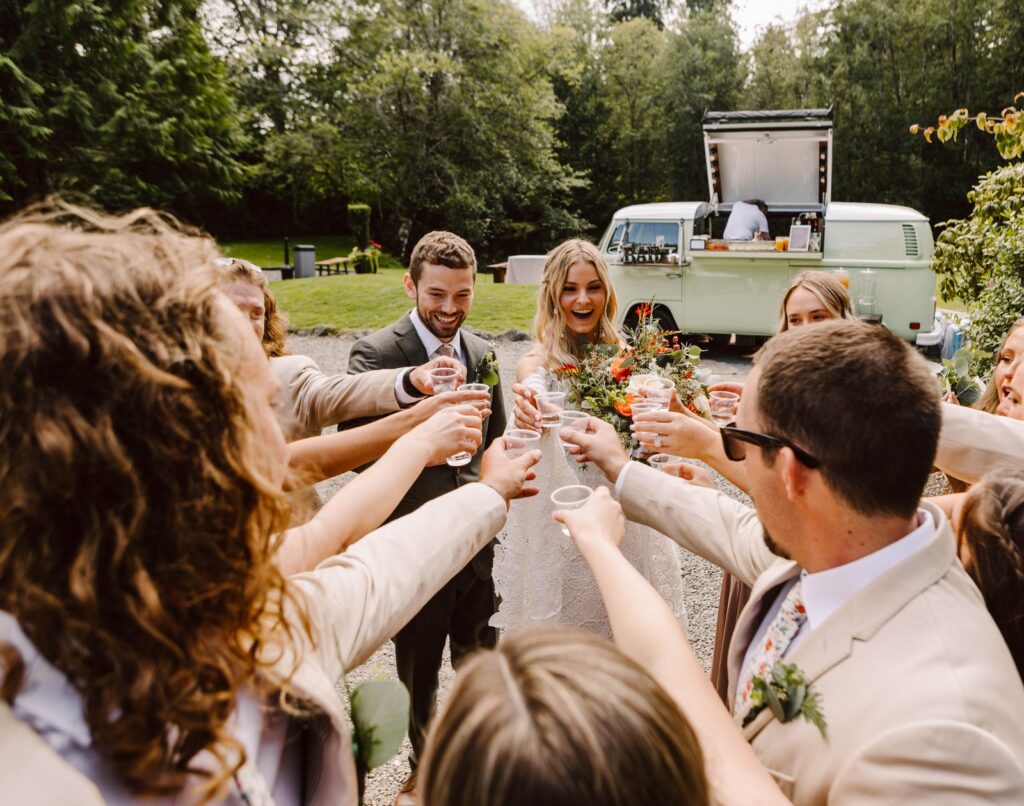 Bride and groom toasting with wedding party outdoors laughing with a vintage vw bus bar and trees in the background