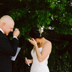 Bride and groom crying while exchanging vows outdoors both wiping tears surrounded by lush green trees