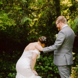 Groom in gray suit comforting emotional bride in white lace dress during an outdoor wedding first look among lush green trees