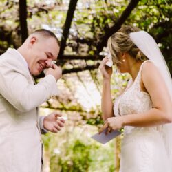 Bride and groom wiping tears and smiling while exchanging vows outdoors beneath a lush green tree canopy