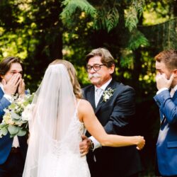 Bride in veil holding bouquet faces emotional father and two tearful groomsmen in blue suits at outdoor wedding ceremony