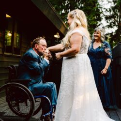 Emotional wedding moment as a man in a wheelchair kisses a brides hand on a wooden deck with a smiling woman in blue watching nearby
