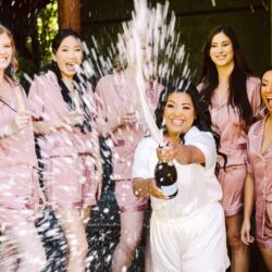 Bride in white popping a champagne bottle while bridesmaids in matching pink satin pajamas cheer and laugh around her