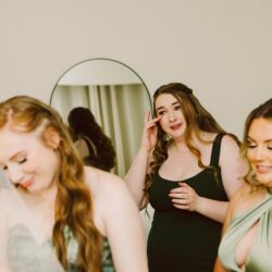 Bridesmaid in black dress wiping away a tear while two other women in sage green dresses smile nearby round mirror in background