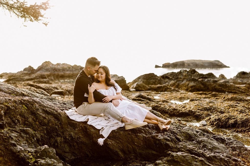 Couple sitting closely on coastal rocks at golden hour man in black embracing woman in white off shoulder dress ocean in background