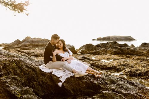 Rosario beach fidalgo island engagement session couple sitting closely on coastal rocks at golden hour man in black embracing woman in white off shoulder dress ocean in background