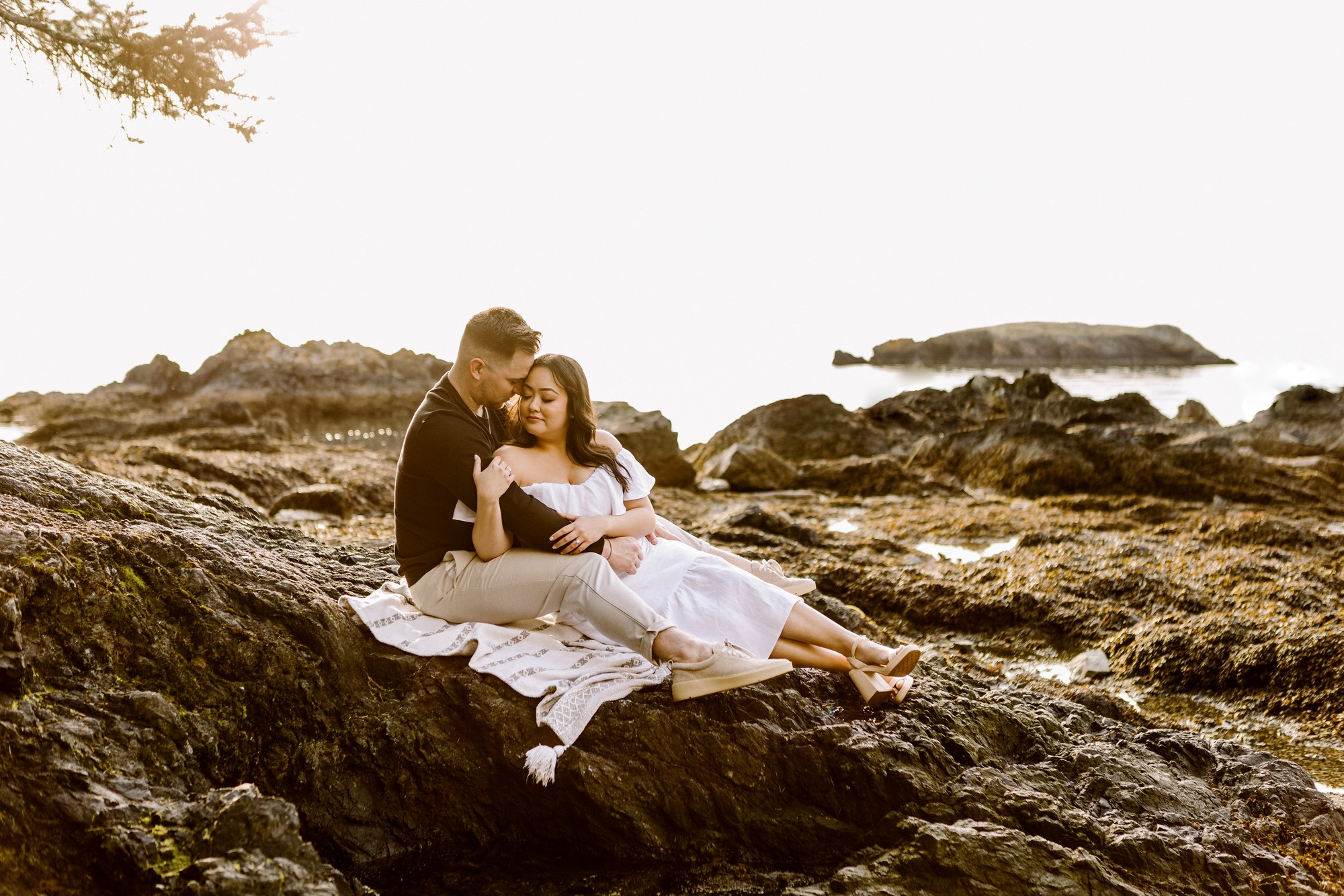 Rosario beach fidalgo island engagement session couple sitting closely on coastal rocks at golden hour man in black embracing woman in white off shoulder dress ocean in background