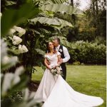 Bride in a white wedding gown holding a bouquet, embraced by her groom in a lush garden with large green banana leaves nearby.
