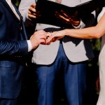 Bride and groom exchange rings outdoors as an officiant reads from a book.