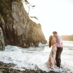Couple embracing and kissing on a rocky beach with waves at their feet and a cliff to the left under a bright sky.
