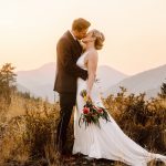 A bride and groom kiss in a mountain meadow at sunset, the bride holding a bouquet and wearing a white gown.