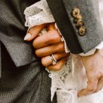 Close-up of two hands clasped together, showing wedding rings, with lace fabric and a gray suit sleeve in the background, suggesting a wedding or formal embrace.