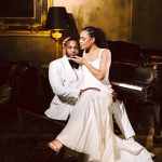 A couple in white formalwear sits by a grand piano in a luxurious room, the woman touching the man's head lovingly.