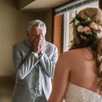 Older man in a light gray suit covers his face with his hands, standing near a window as a bride in a wedding dress faces him from behind.