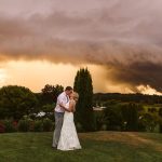 Couple in wedding attire embracing and kissing on a grassy hill as a dramatic orange sunset and dark storm clouds loom in the background.