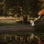Bride and groom in wedding attire walk along a path by a pond in a park with a wooden gazebo nearby.