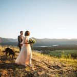 Bride in a flowing white gown holds a bouquet as she walks on a hilltop at sunset, accompanied by a groom in a suit and a dog with a valley and mountains in the distance