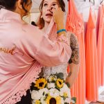 Bridal stylist in a pink robe adjusts the bride's makeup as the bride holds a bouquet of sunflowers and white flowers in a pastel dress shop.