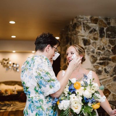 Snohomish Wedding Photographer GSquared Weddings Two women joyfully converse in a warmly lit room. One wears glasses and a floral dress, while the other, in a white dress holding a bouquet of flowers, seems to be celebrating her wedding. The stone wall behind them adds to the charm of this special occasion. Serving Seattle, Snohomish and Orlando
