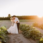 A couple kissing in a white wedding dress and black hat in a rural field at sunset, embracing among leafy plants.