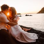 Couple embracing and kissing on rocky shoreline at sunset with a sun flare between them over the calm sea.