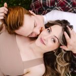 Couple cuddling on grass, close-up of a man with red hair kissing or nuzzling a woman with blue eyes and light makeup.