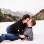 Couple kissing in the snow with mountain scenery in the background.