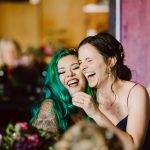 Two women laughing and embracing at a party, one with teal hair and a tattooed arm visible in the foreground.