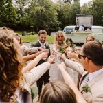 Bride and groom toasting with guests outdoors, bouquets in view, next to a vintage camper bar in a garden setting.