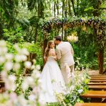Bride and groom embrace under a colorful flower arch in a forested outdoor wedding setting beneath trees and greenery, with wooden benches nearby.