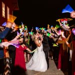 Bride and groom kiss in the center as guests cheer with colorful pennants at night.