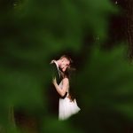 Couple kissing in a forest clearing, framed by tree trunks and lush green leaves around them.