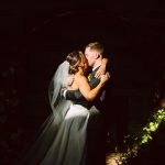 Bride and groom share a kiss while embracing, the bride in a white gown and veil, against a dark background.